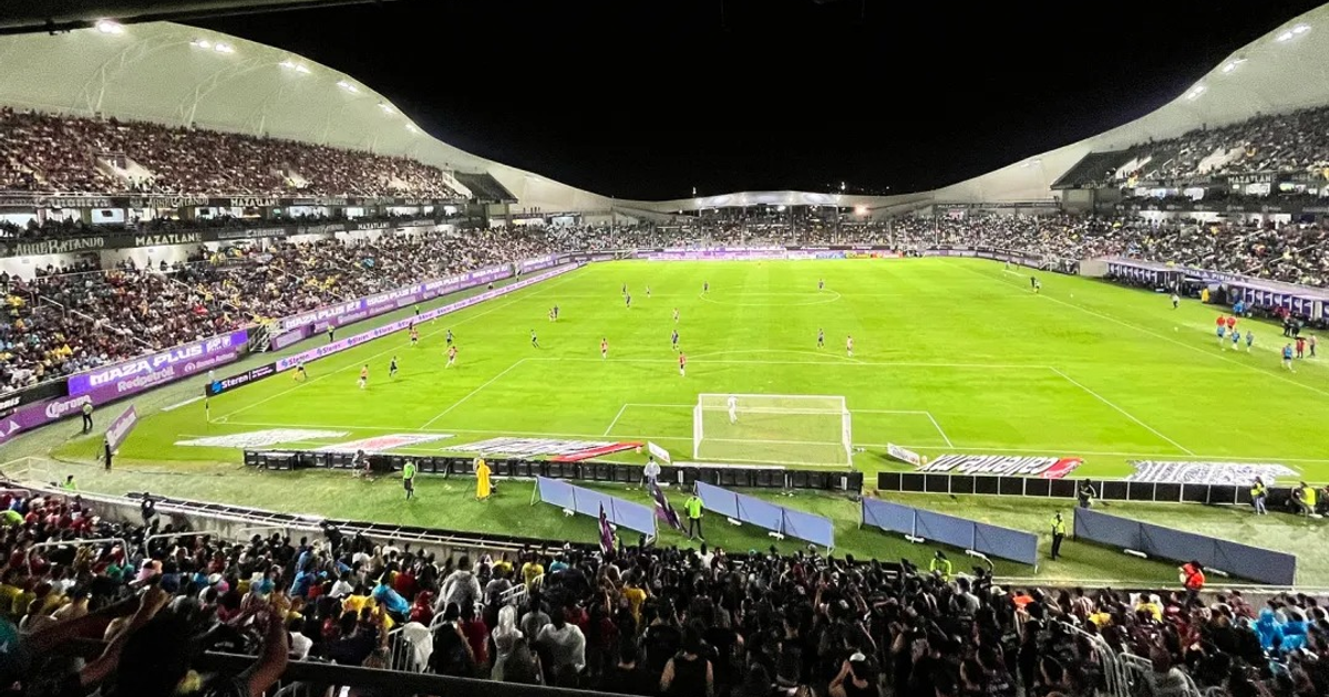 Estadio de fútbol lleno de espectadores durante un partido nocturno, con jugadores en el campo y aficionados animando desde las gradas.