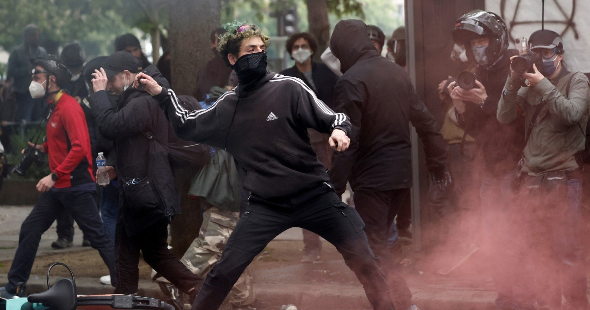 Manifestante enmascarado lanza objeto durante protestas del 1 de mayo en Francia, con humo rosa en el aire.