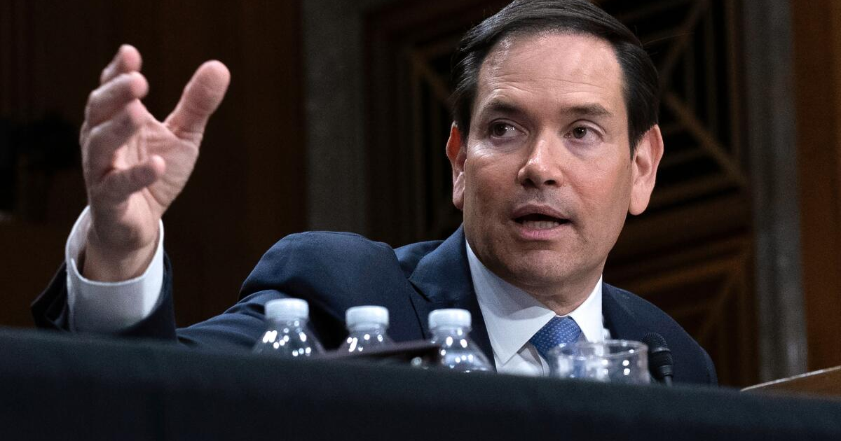 Persona gesticulando en una audiencia formal con botellas de agua y micrófono en la mesa.