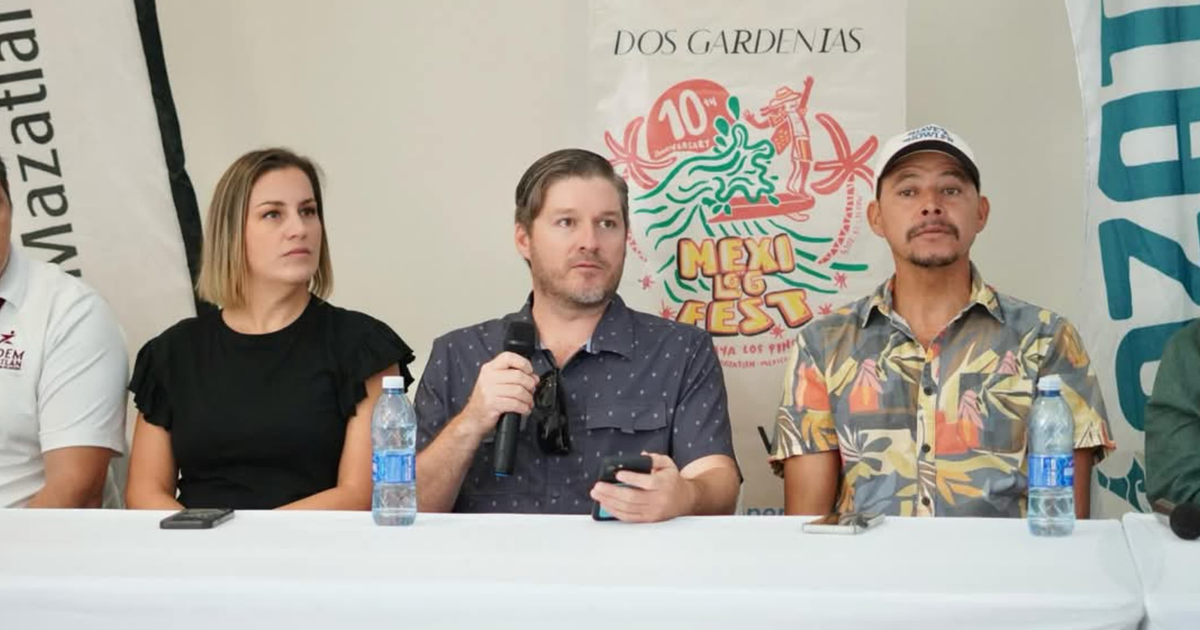 Personas reunidas en una mesa durante el Mexilog Fest en Mazatlán, con un cartel del evento en el fondo.