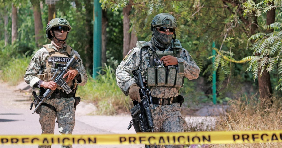 Dos soldados con uniformes tácticos y armas en un entorno boscoso con cinta de precaución amarilla.