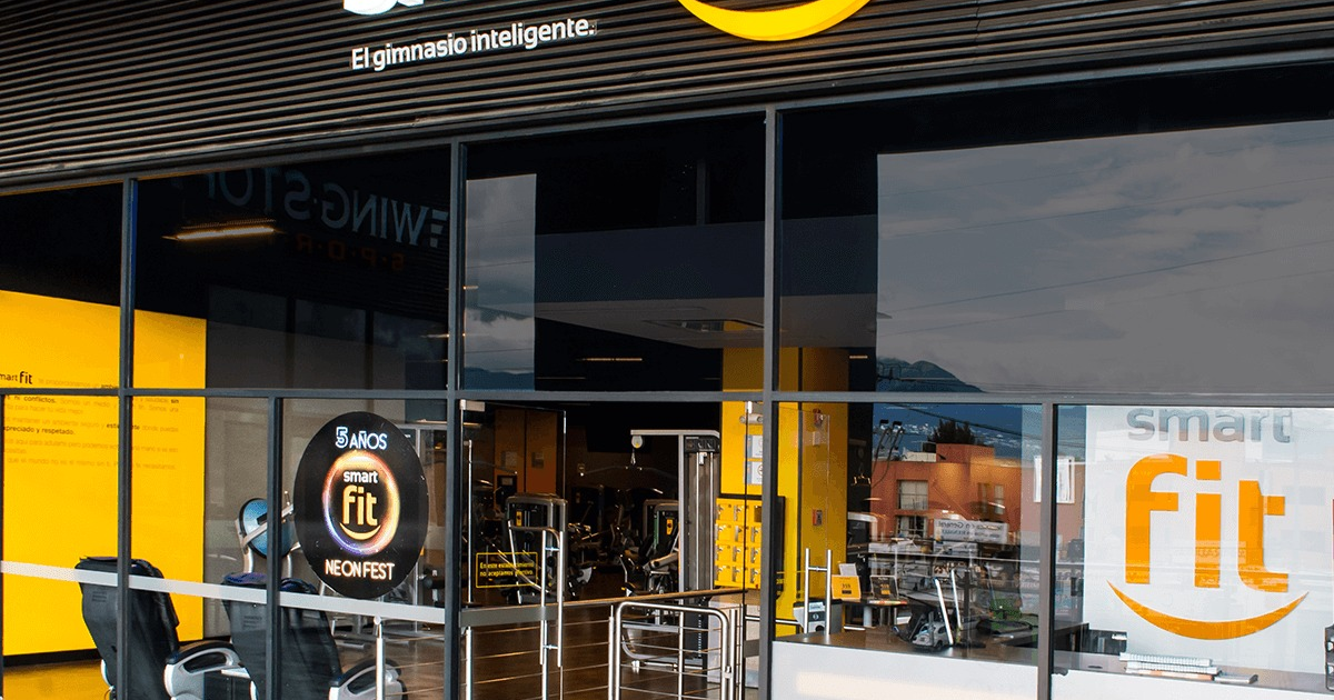 Entrada del gimnasio Smart Fit con logo y lema, decorado amarillo y vista interior de máquinas de ejercicio.
