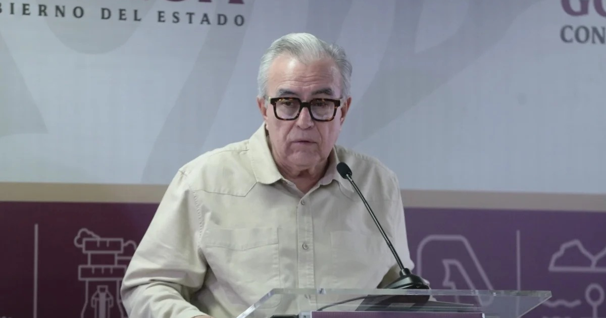 Hombre de cabello canoso y gafas oscuras hablando en un podio con micrófono, fondo con gráficos de gobierno estatal.