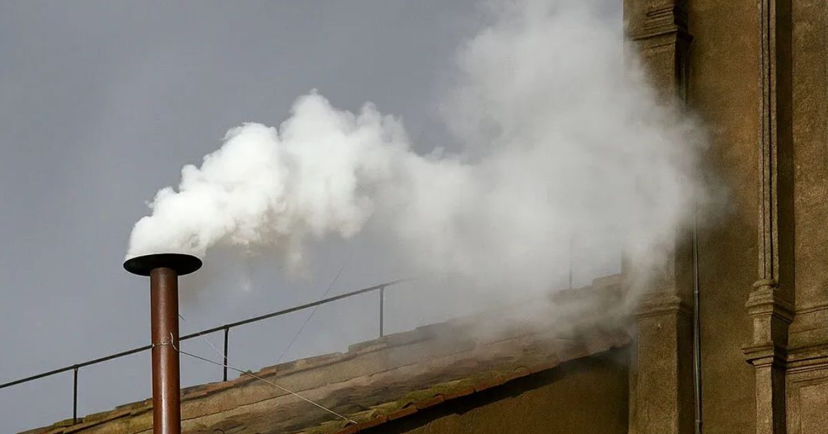 Chimenea emitiendo humo blanco, señal de elección papal, con cielo nublado y edificios de fondo.