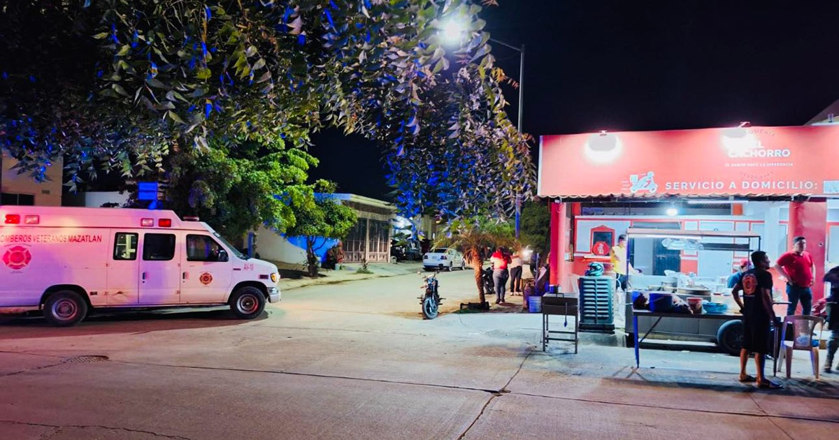 Ambulancia con luces encendidas en calle nocturna frente a 'El Chorro', un establecimiento de comida, con personas en la calle y sombras de árboles.