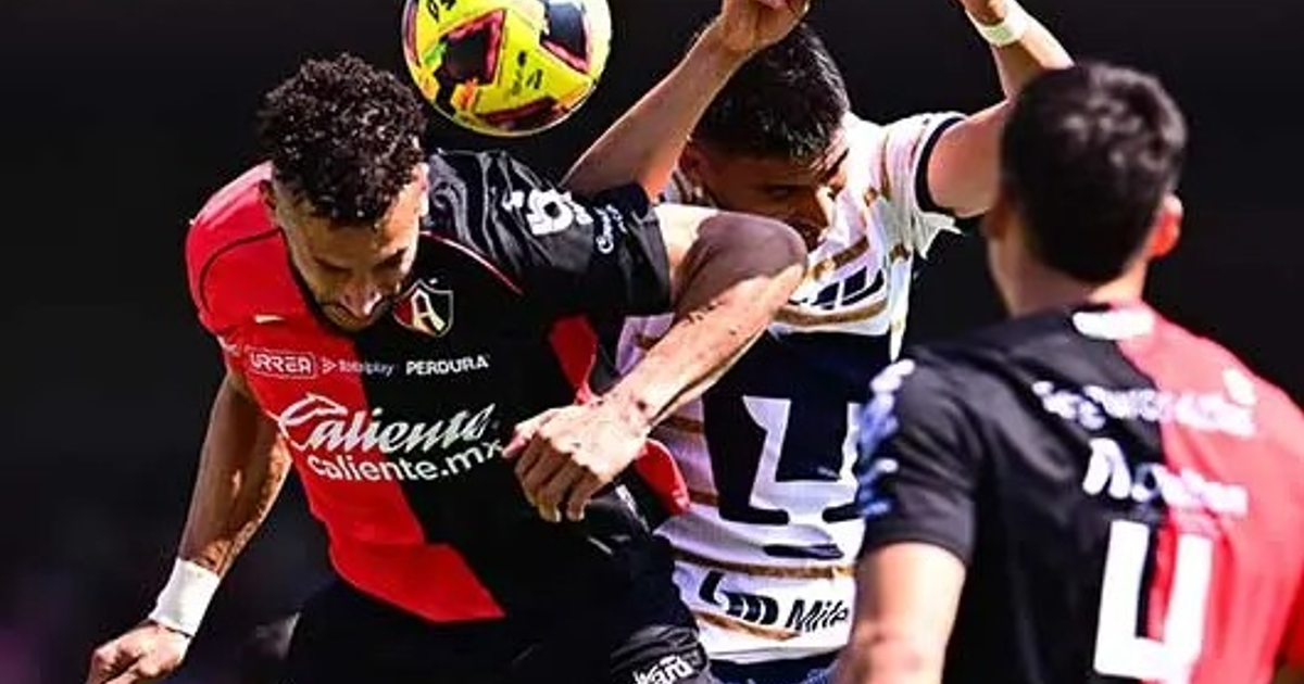 Dos jugadores de fútbol disputan el balón en el aire, uno con camiseta roja y negra y otro con camiseta blanca y azul, en un estadio.