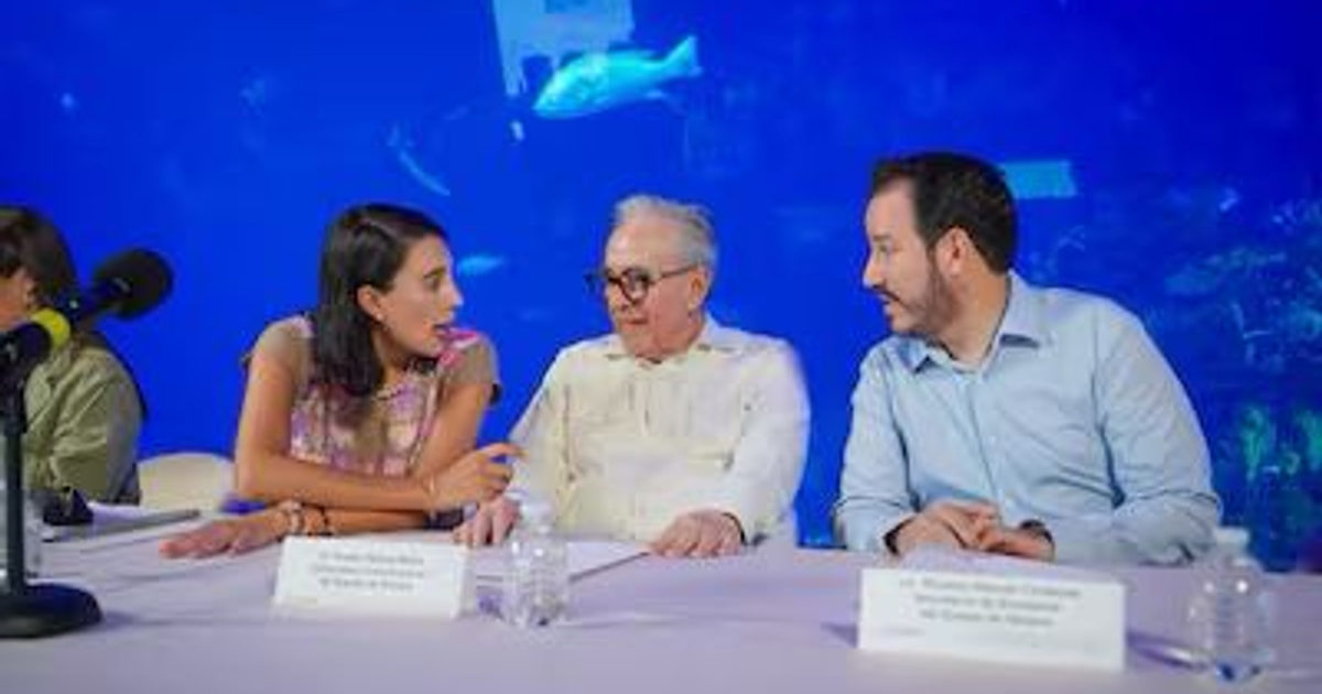Tres personas conversando en una mesa con un micrófono, fondo azul de acuario, relacionado con Reverb by Hard Rock en Mazatlán.