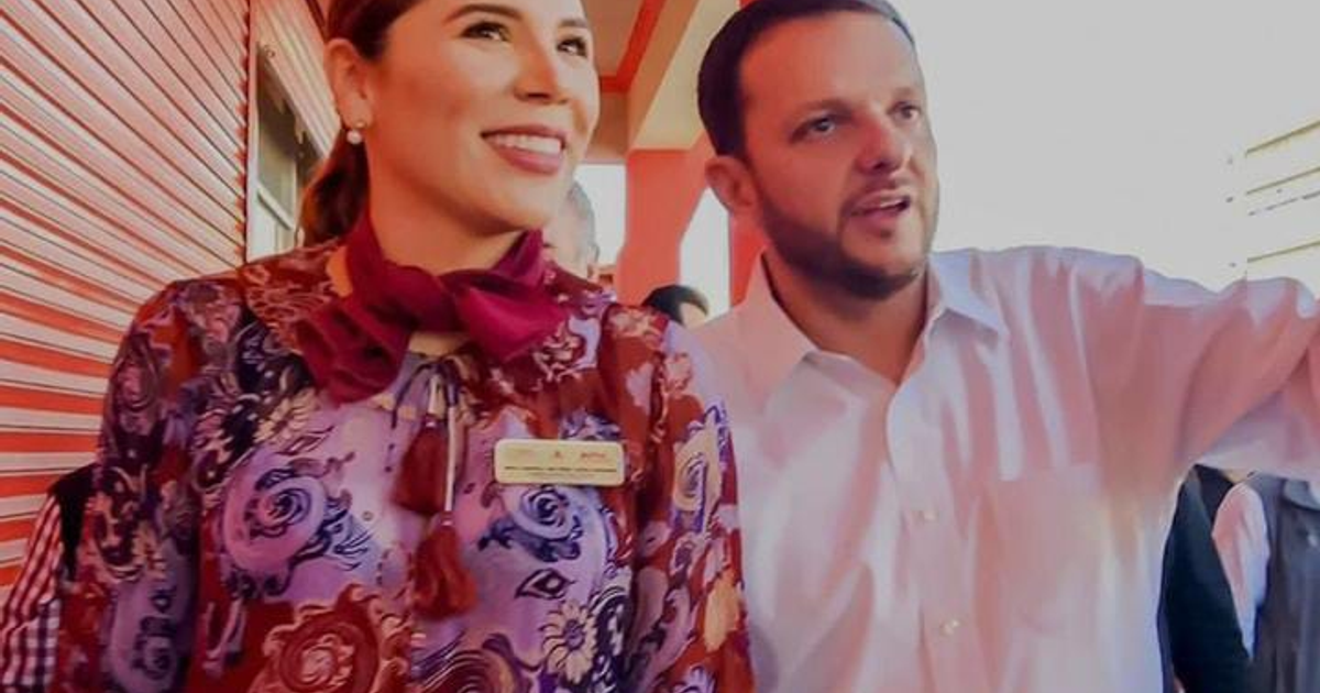 Dos personas sonriendo y conversando en un evento social con fondo de colores cálidos.