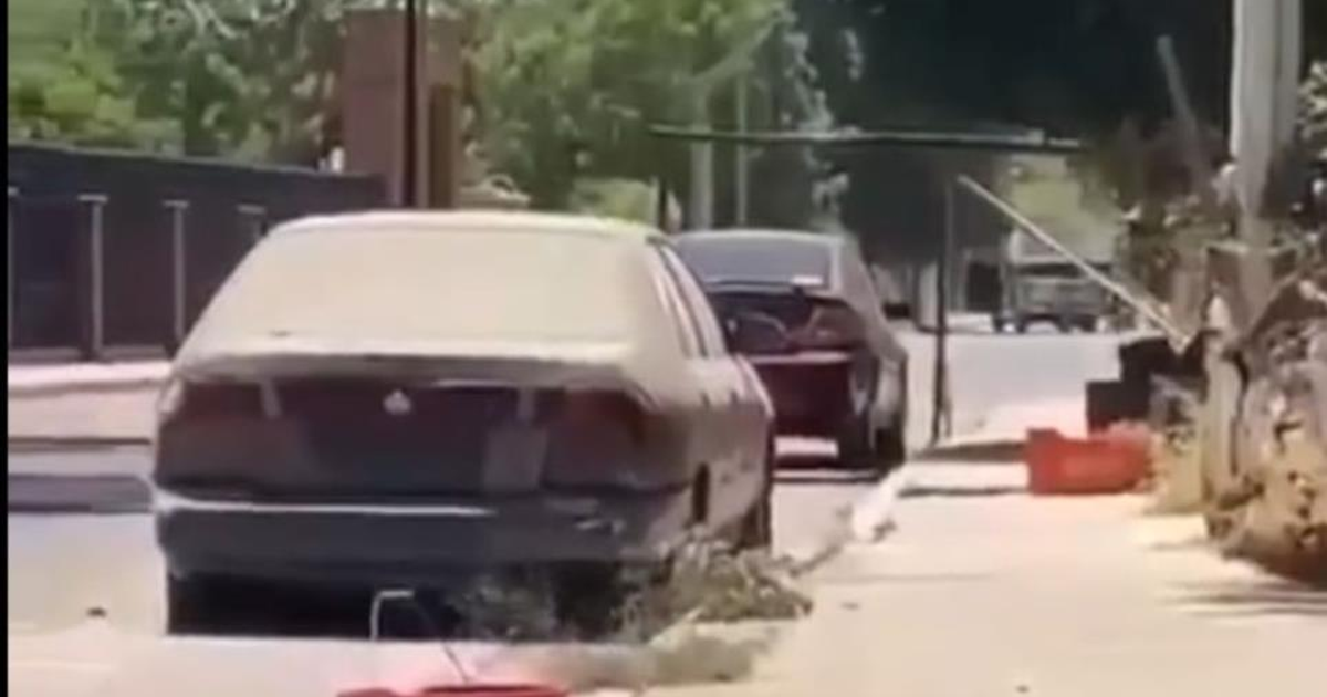 Dos vehículos estacionados en una calle urbana de Mocorito, Sinaloa, con vegetación y un objeto naranja en la acera.