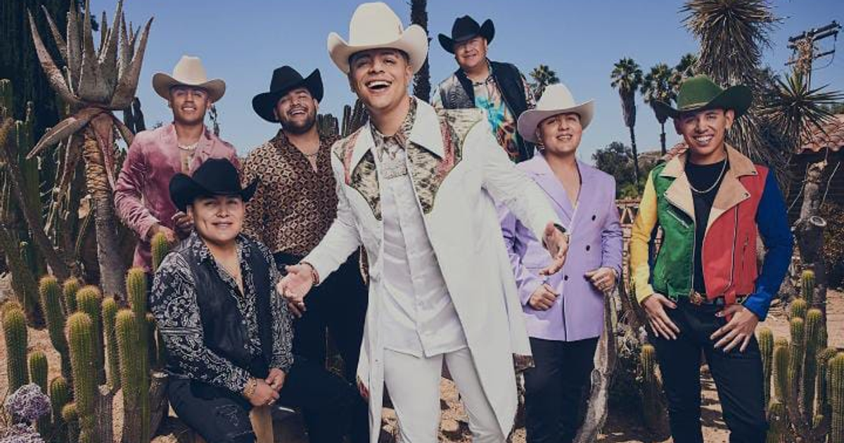 Grupo Firme posando en el desierto con trajes coloridos y sombreros, rodeados de cactus y palmeras.