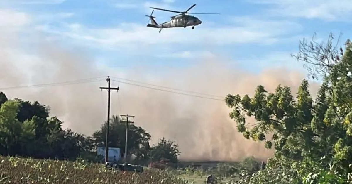 Helicóptero militar volando sobre área rural con nube de polvo al fondo