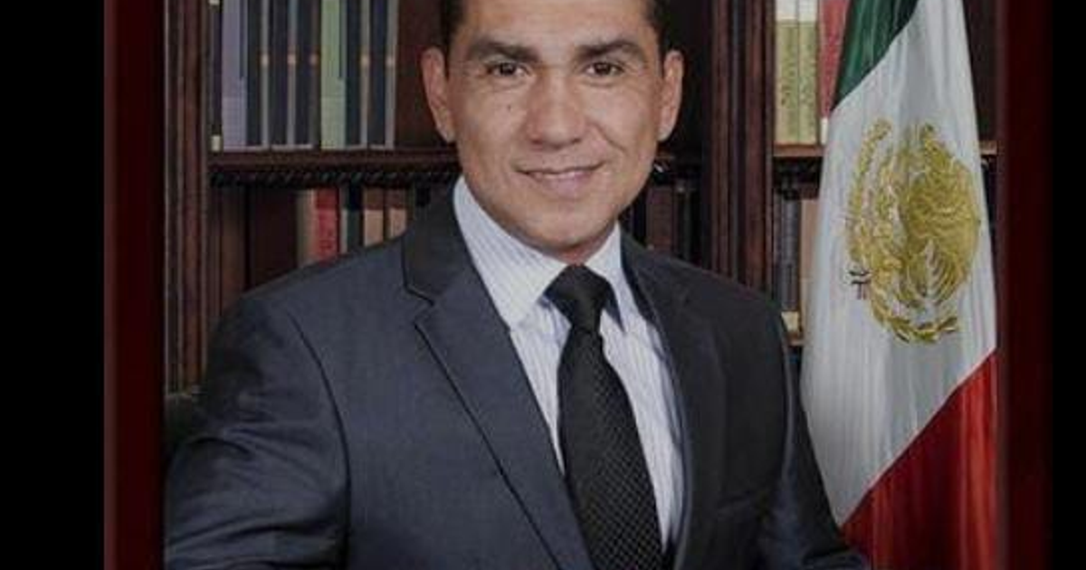 Hombre con traje oscuro sonriendo frente a biblioteca y bandera de México