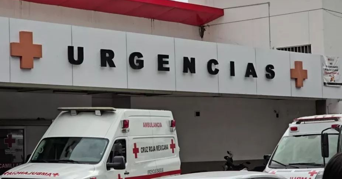 Letrero de urgencias con cruz roja y ambulancias estacionadas frente a un edificio de emergencias médicas.
