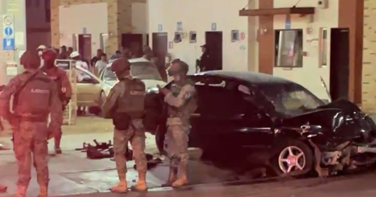 Auto negro dañado en escena de balacera nocturna en Culiacán, rodeado de seguridad y espectadores.