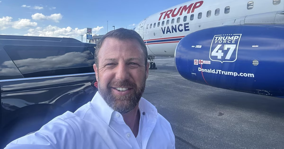 Persona sonriente tomando selfie frente a avión 'TRUMP VANCE' y 'TRUMP FORCE 47', vehículo negro al lado, cielo despejado.