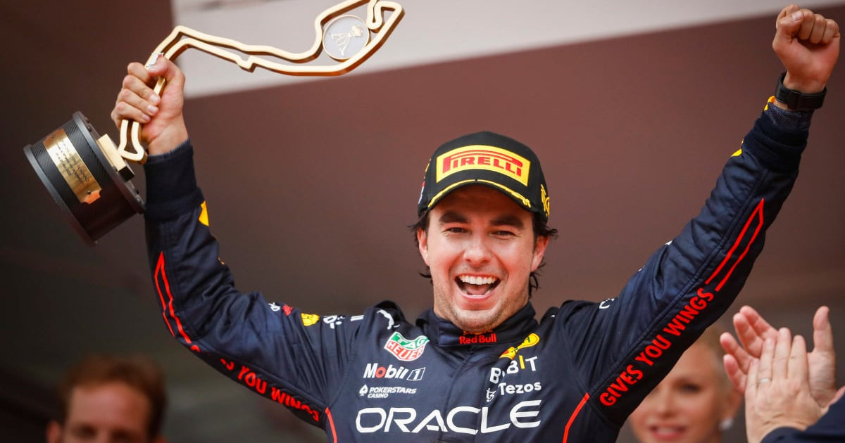 Piloto de Fórmula 1 celebrando en el podio con un trofeo, vestido con traje negro y rojo.