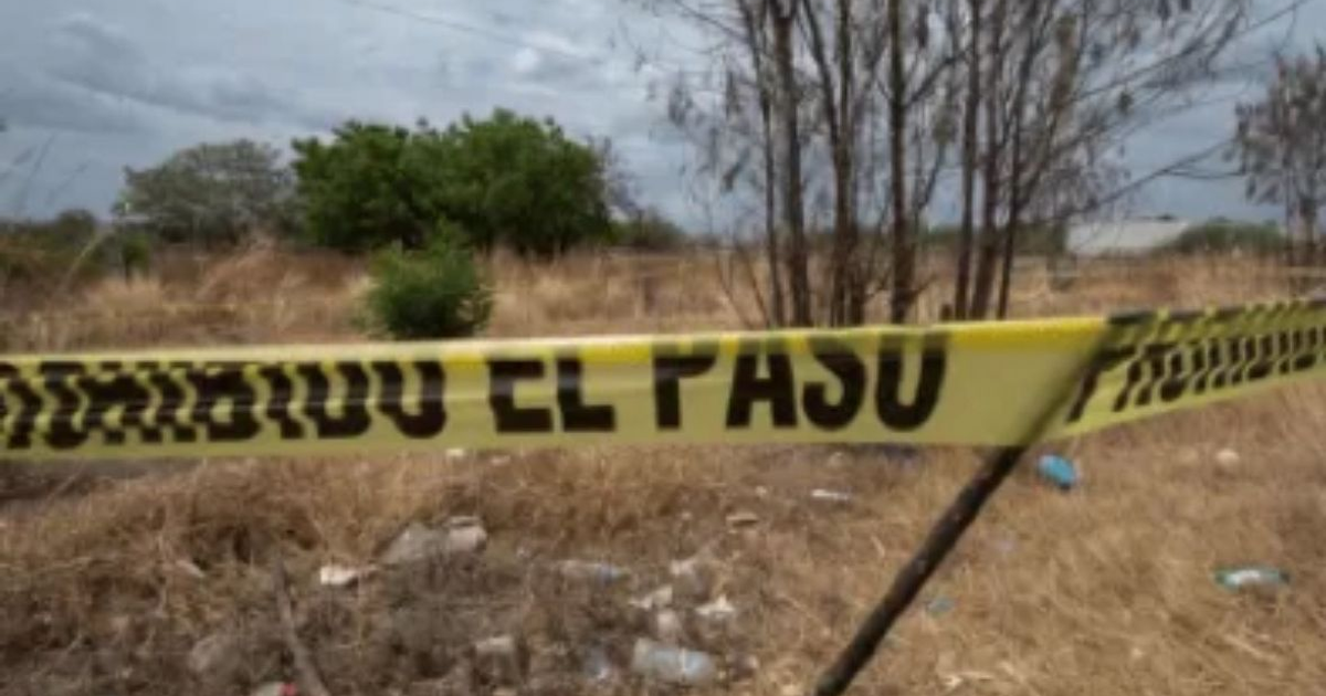 Cinta amarilla de 'PROHIBIDO EL PASO' en terreno descuidado con vegetación y cielo nublado.