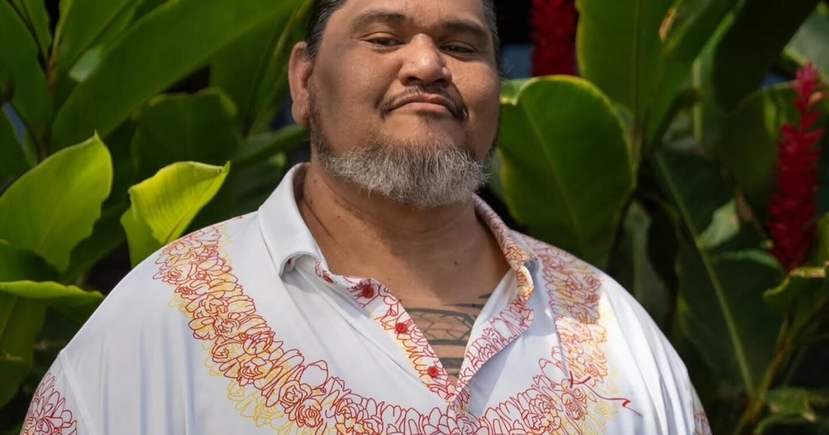 David Hekili Kenui Bell de pie frente a un fondo de plantas verdes, con camisa blanca y diseño colorido, en un ambiente tropical.