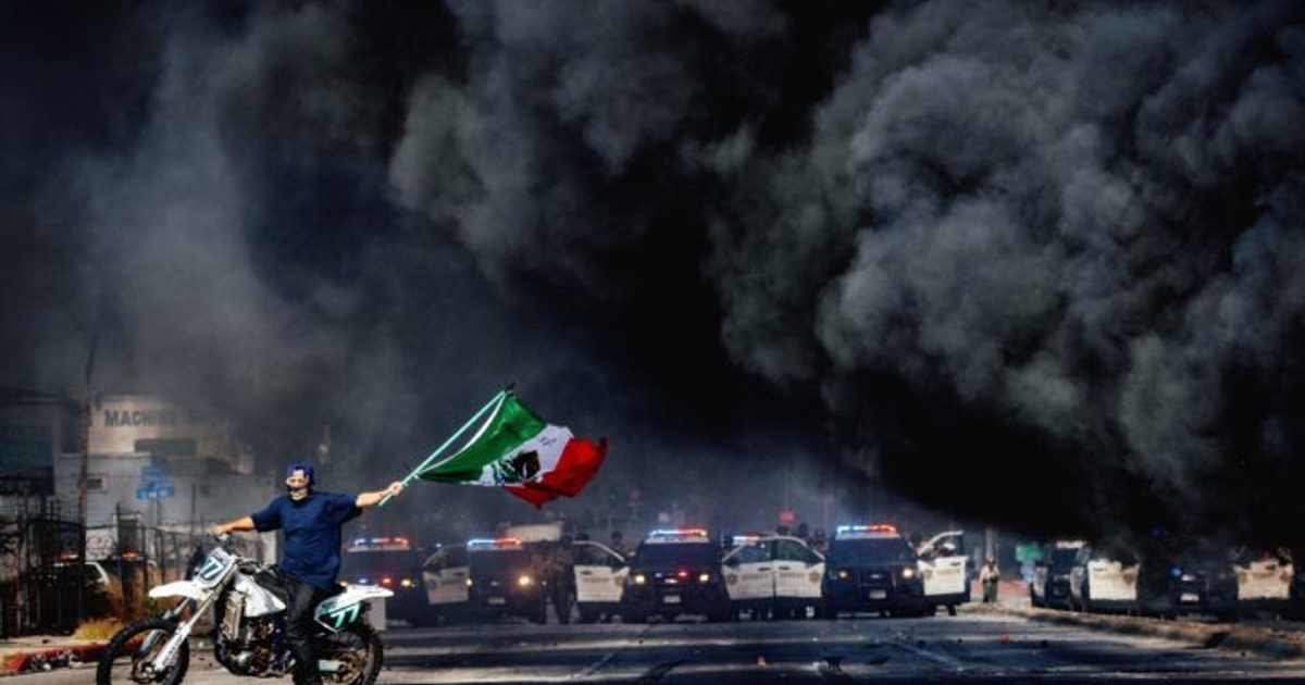 Hombre en motocicleta con bandera mexicana y humo negro en el fondo, rodeado de vehículos policiales.
