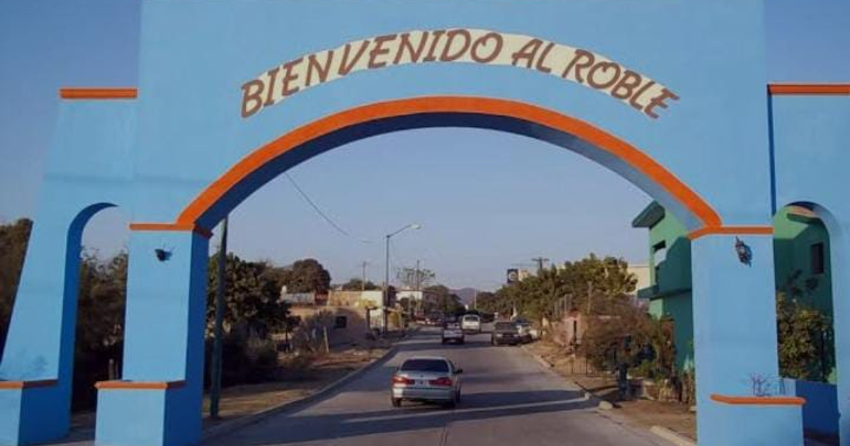 Arco de entrada azul y naranja de El Roble sobre carretera con vehículos, fondo de edificaciones y árboles.