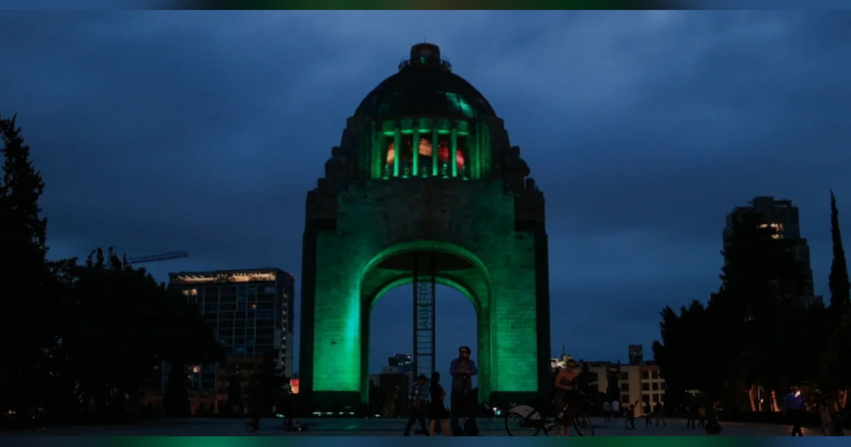 Monumento iluminado en verde en el Centro Histórico de la Ciudad de México, con personas caminando y edificios modernos al fondo.