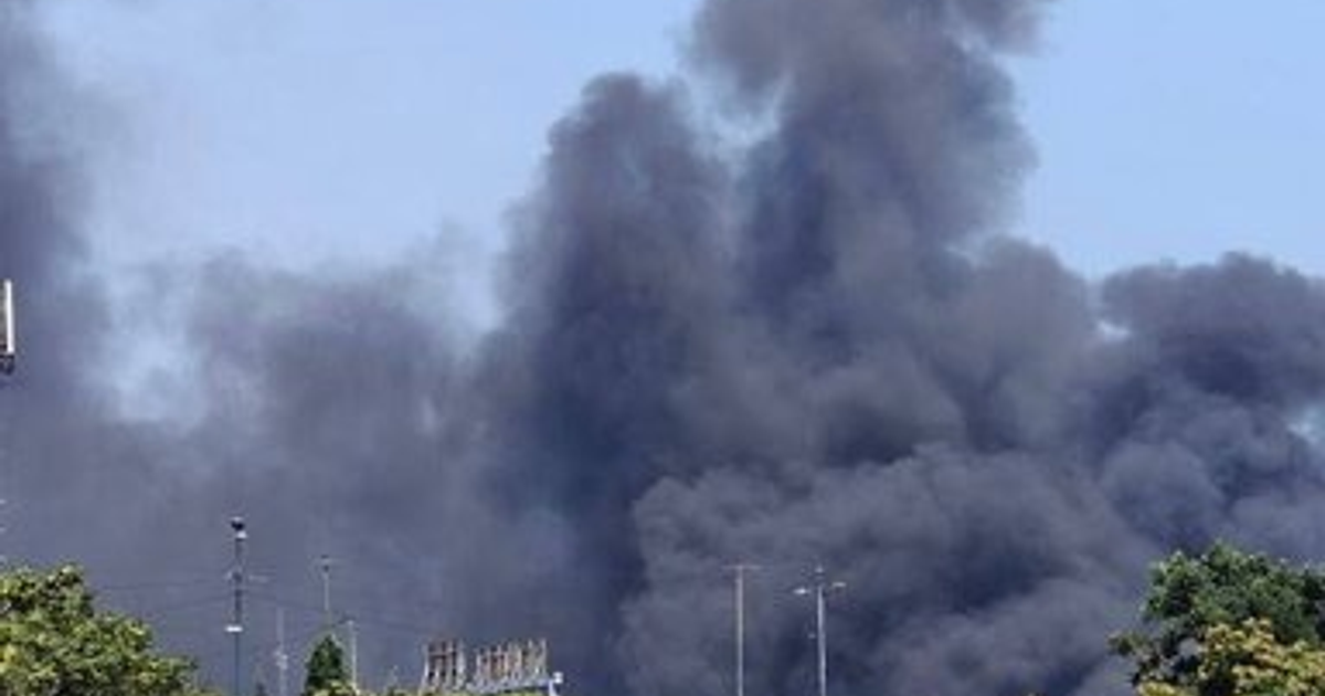 Columna de humo negro en el cielo azul con estructuras y árboles en el fondo.