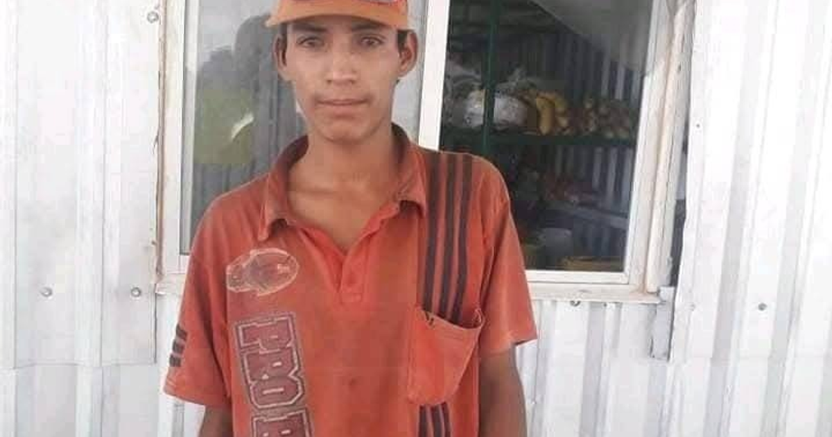 Hombre con camisa naranja y gorro frente a ventana de negocio con frutas.