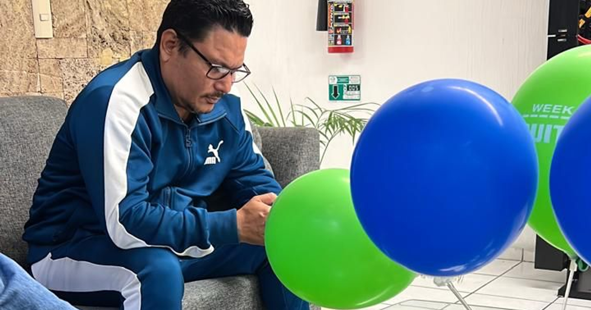 Hombre en sofá con chaqueta azul, rodeado de globos de colores, mirando su teléfono móvil.