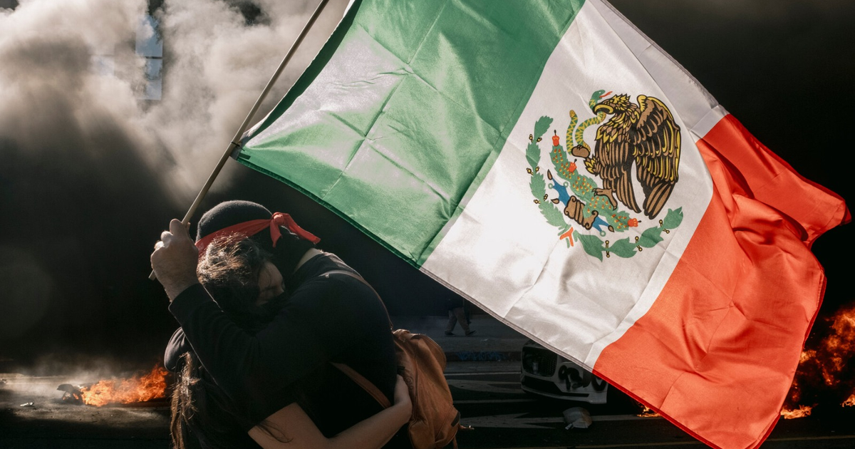 Dos personas abrazadas sosteniendo una bandera de México en una protesta con humo y llamas de fondo.