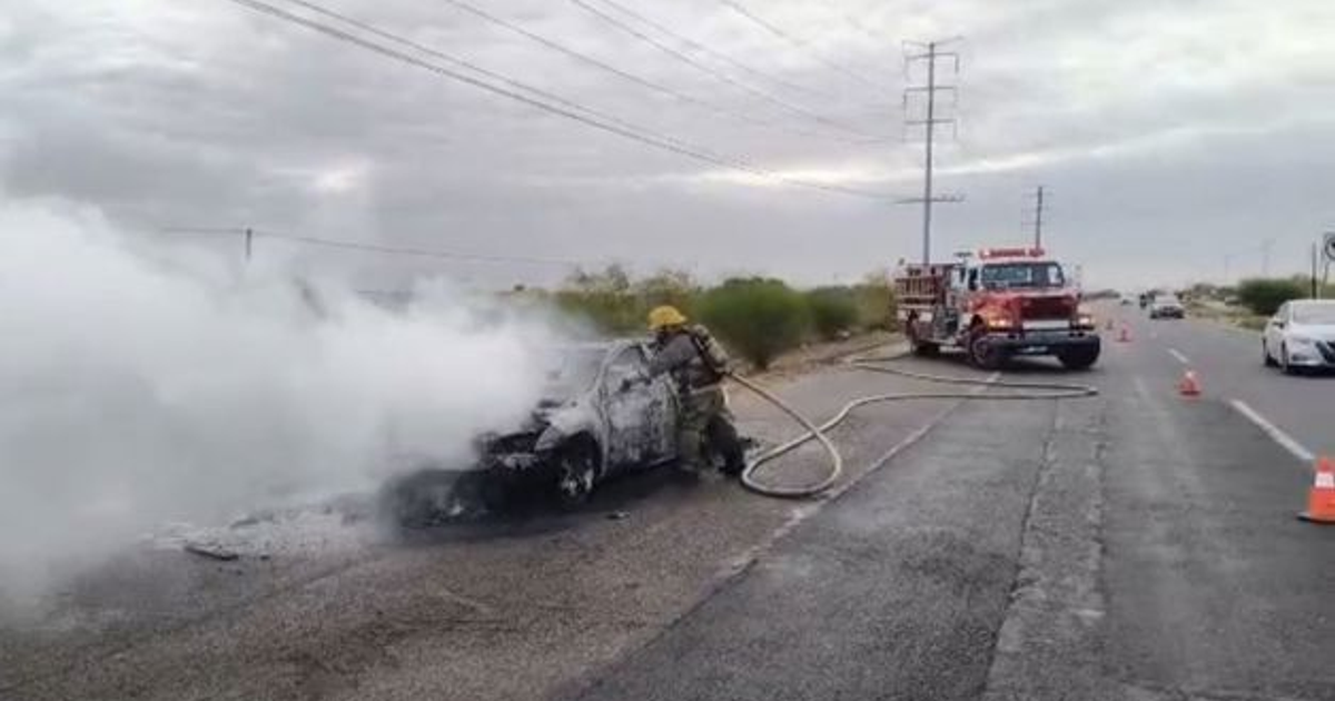 Bombero apaga incendio de vehículo en carretera con manguera, humo denso y camión de bomberos al fondo.