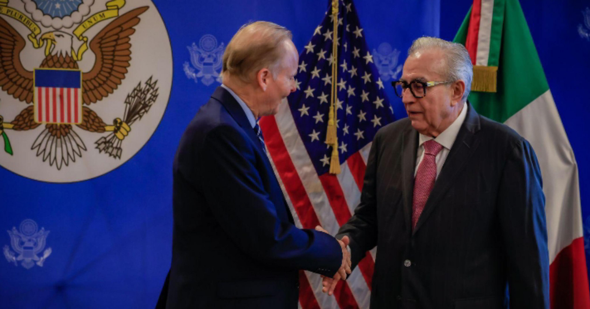Dos hombres estrechándose la mano en un evento diplomático con banderas de Estados Unidos y México al fondo.