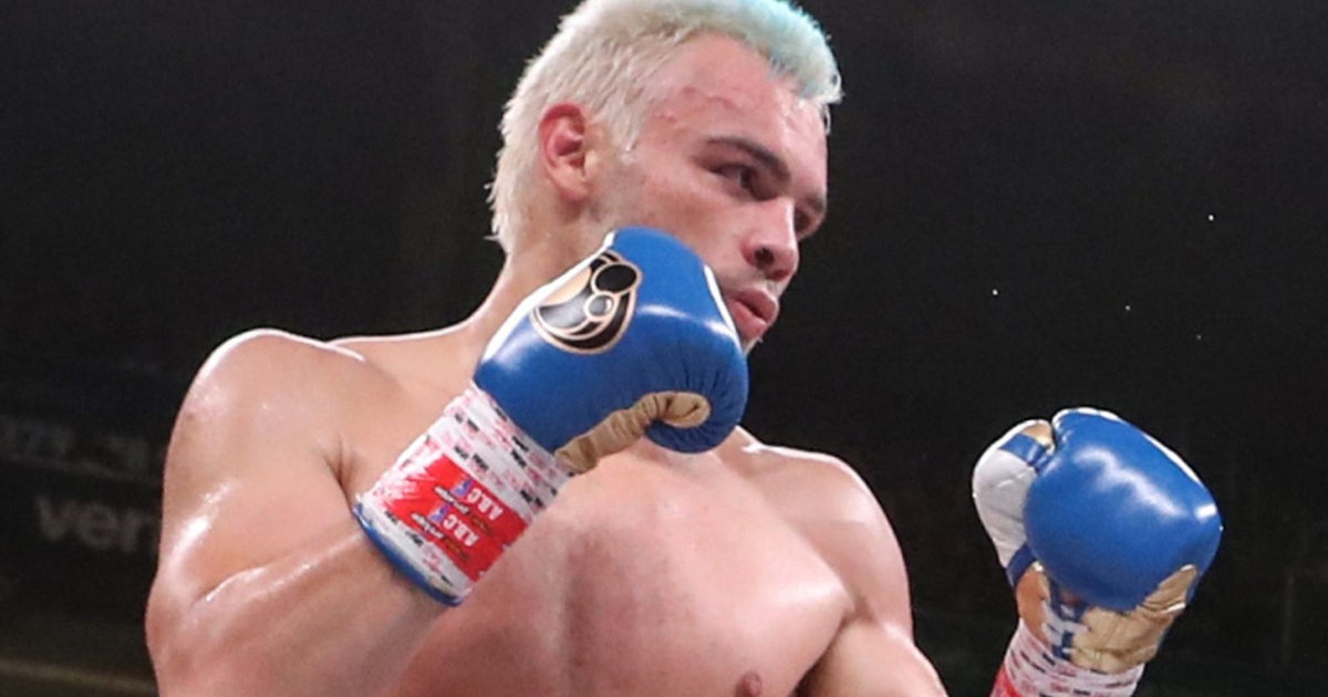 Boxeador rubio con guantes azules en el ring, concentrado y en postura defensiva.