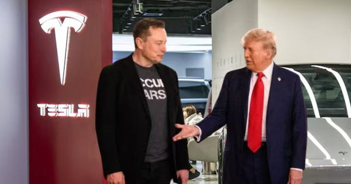 Dos personas conversan en una instalación de Tesla, con el logotipo de Tesla visible en la pared y vehículos en el fondo.