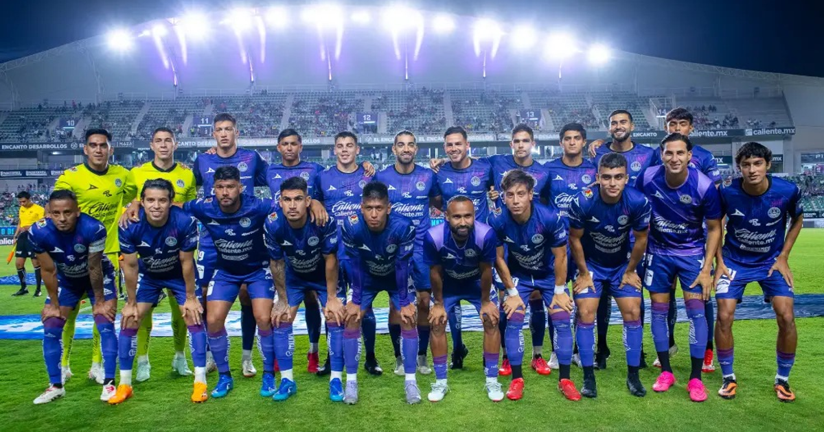 Equipo de Mazatlán FC posando en el campo con uniformes azules y portero en amarillo, estadio iluminado de fondo.