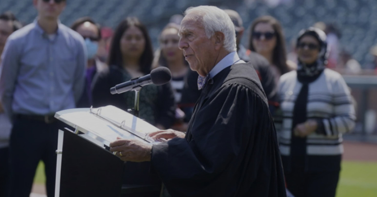 Hombre mayor en toga hablando en micrófono durante evento al aire libre