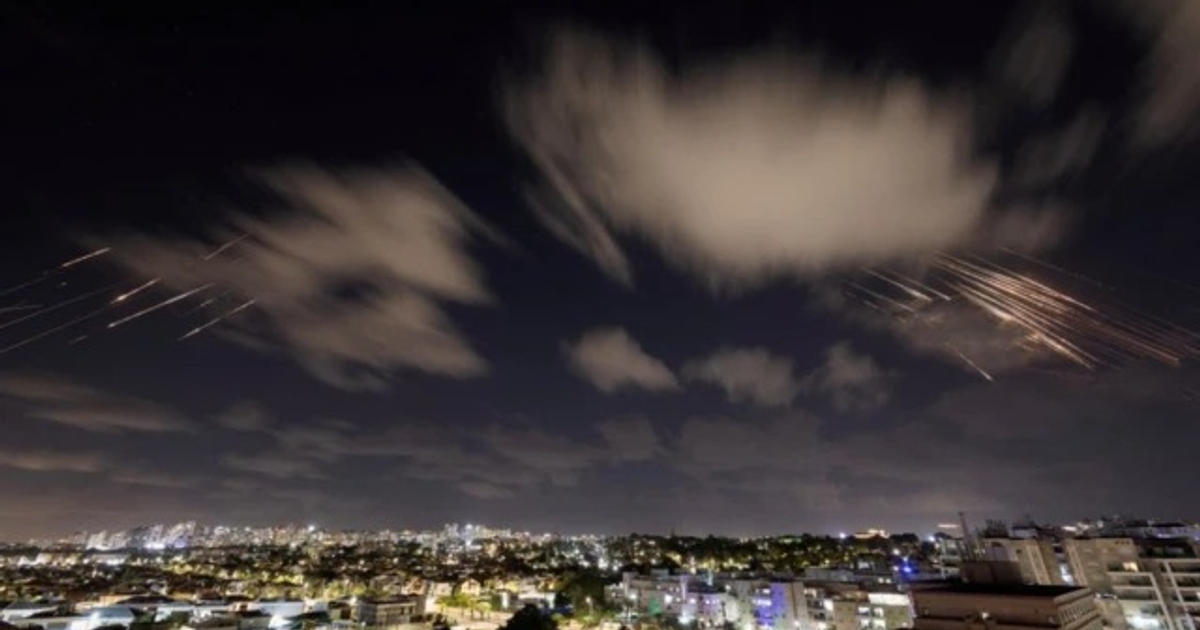 Paisaje urbano nocturno con luces de la ciudad y trazas luminosas en el cielo, posiblemente proyectiles o fuegos artificiales.