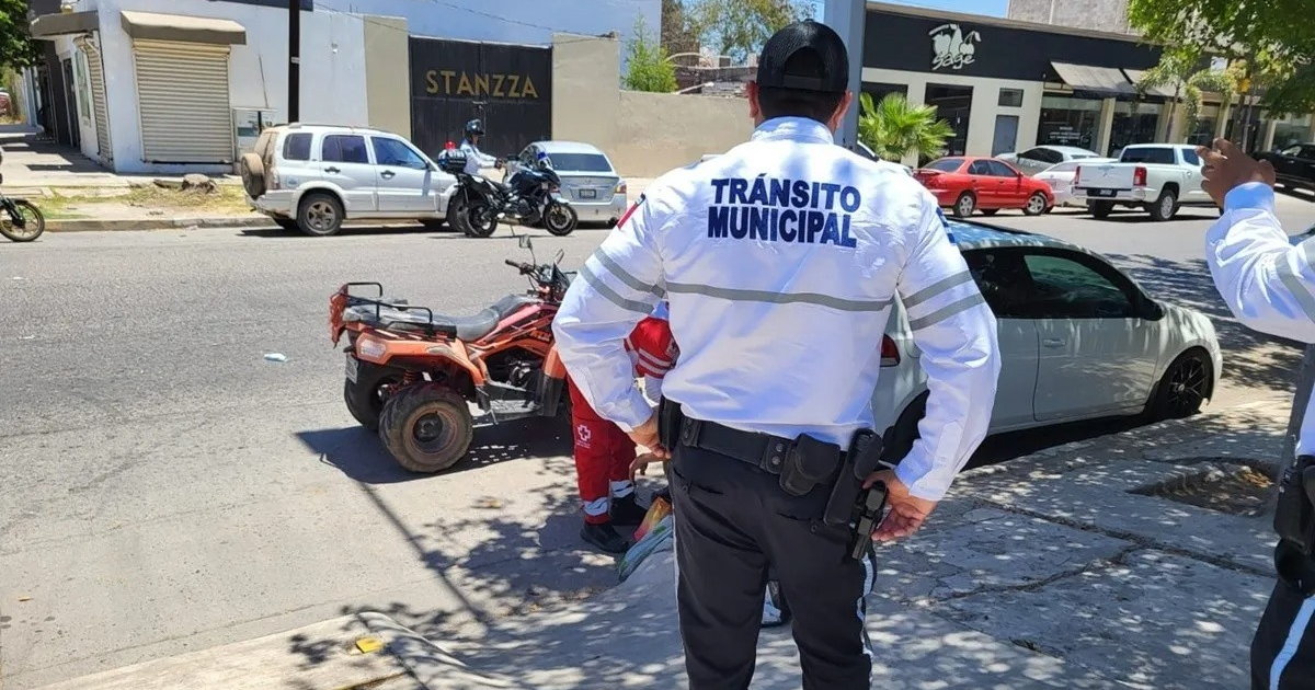 Oficial de tránsito de espaldas junto a un vehículo rojo en una calle de Los Mochis, con un edificio y árboles al fondo.