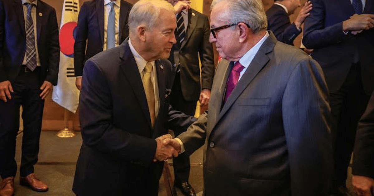 Dos hombres estrechándose la mano en un evento oficial con una bandera de Corea del Sur al fondo.