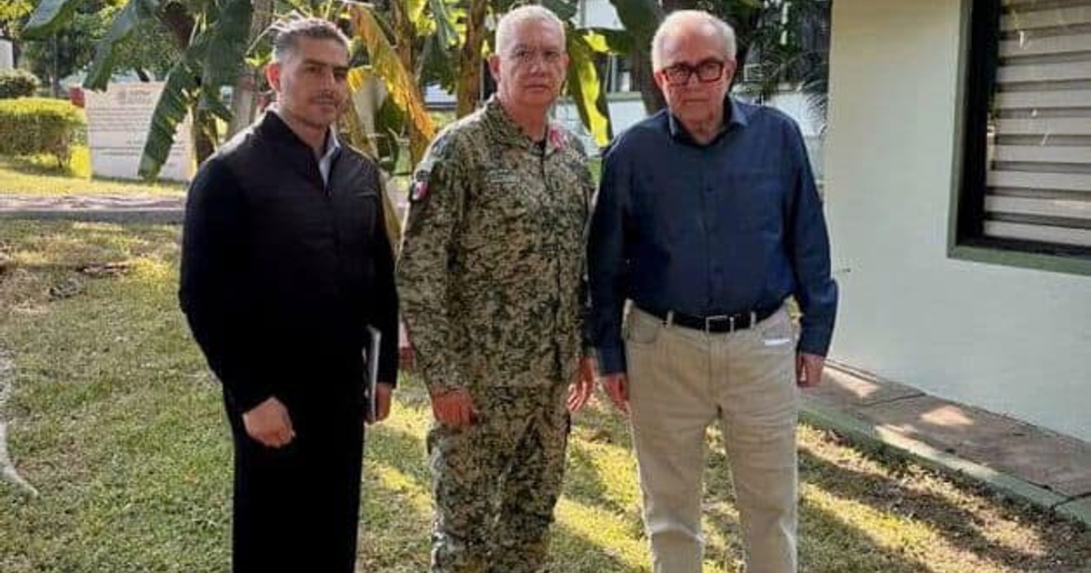 Tres personas en un entorno al aire libre, una con uniforme militar y dos con ropa casual, junto a una pared blanca y plantas.