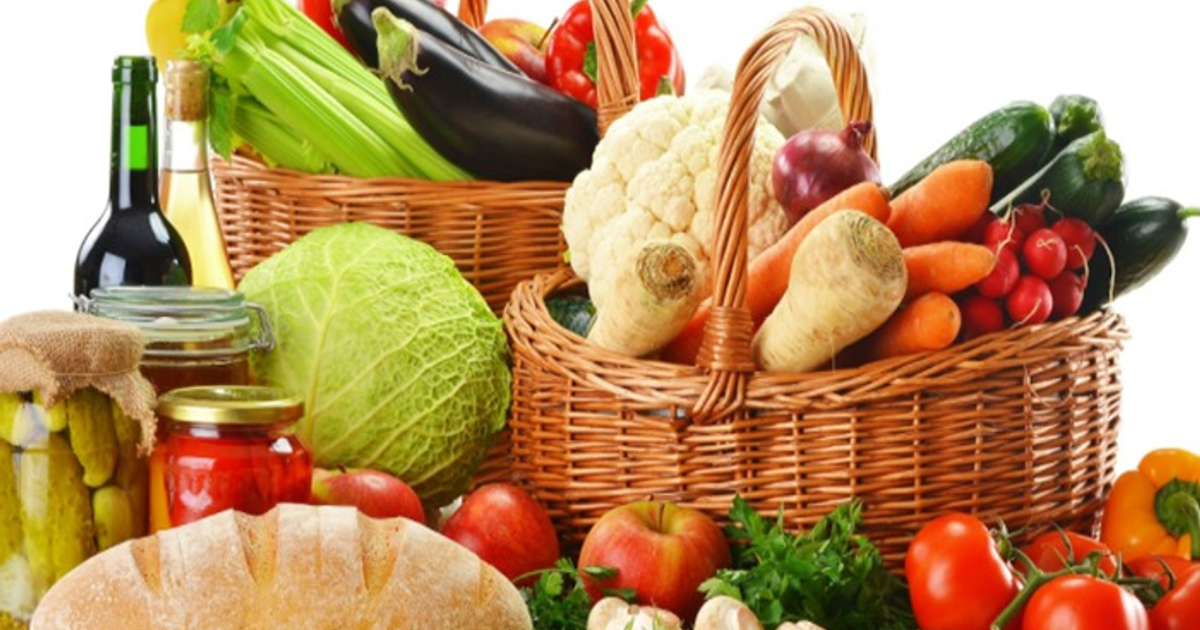 Cestas de mimbre con frutas y verduras frescas, incluyendo col, zanahorias y manzanas, junto a botellas y tarros.