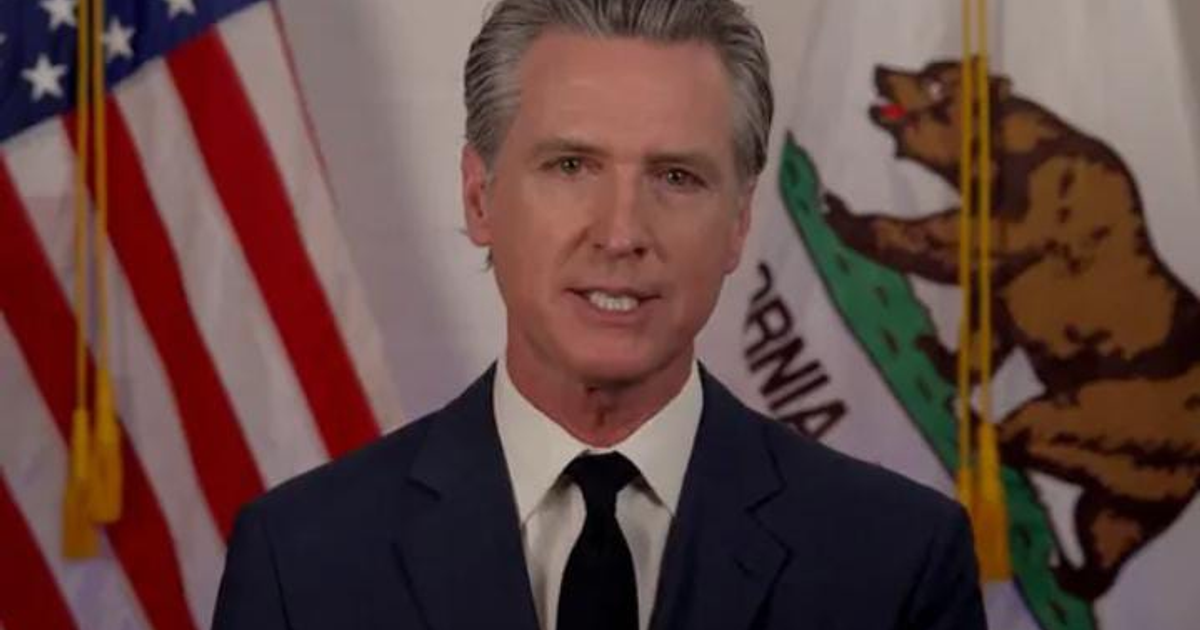 Gavin Newsom hablando sobre redadas migratorias en Los Ángeles con banderas de EE.UU. y California de fondo.