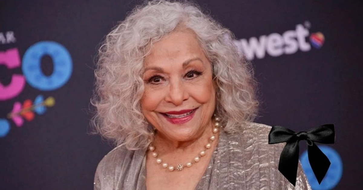 Mujer con cabello rizado y canoso, blusa brillante y collar de perlas, fondo colorido y moño negro.