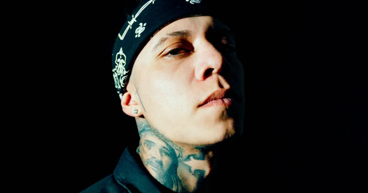 Retrato de Santa Fe Klan con pañuelo en la cabeza y tatuajes visibles en el cuello, fondo oscuro.