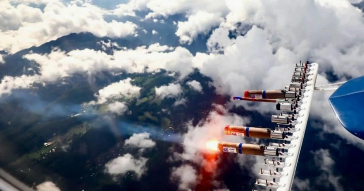 Avión en vuelo sobre montañas nubladas, mostrando dispositivos de siembra de nubes en el ala.