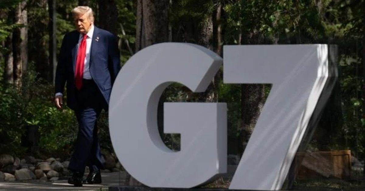 Persona con traje y corbata roja caminando junto a letrero 'G7' en un entorno natural