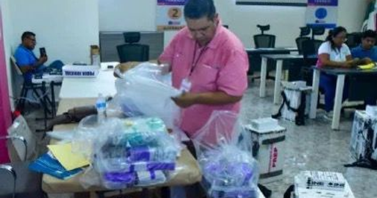 Hombre revisando bolsas en evento electoral con mesas y personas al fondo