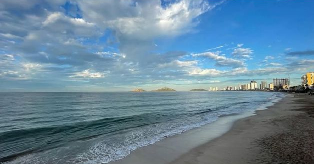 Playa tranquila en Mazatlán con edificios y colinas al fondo, cielo parcialmente nublado.