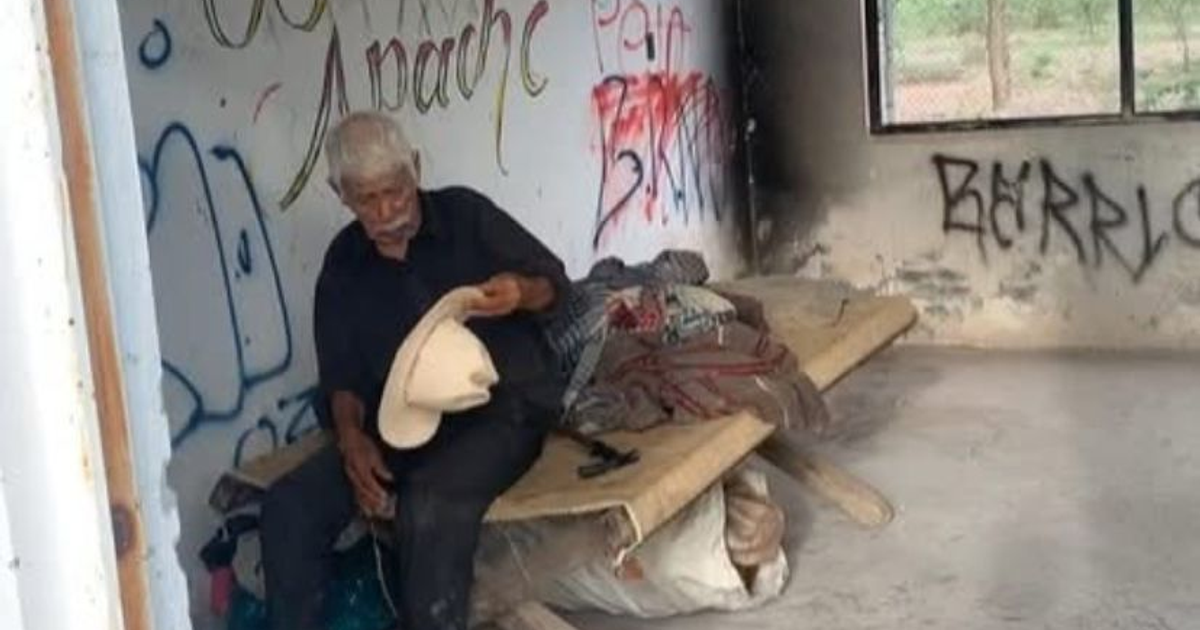 Hombre mayor en habitación abandonada con grafitis y desorden, sosteniendo un sombrero.
