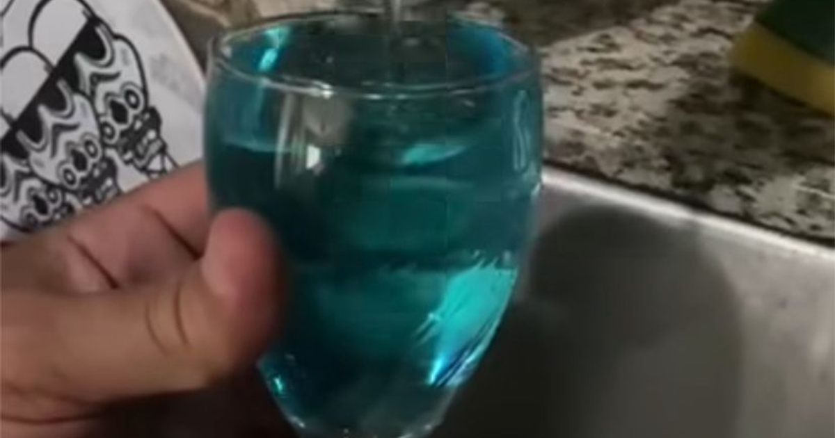 Vaso de cristal con agua azul sostenido por una mano, sobre una encimera clara, con una esponja amarilla al lado.