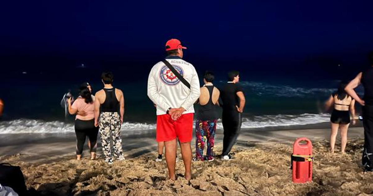 Personas en la playa de Mazatlán durante la noche, con un salvavidas en primer plano, observando el mar.